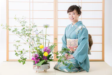 A woman wearing a blue kimono sits on a tatami mat beside a traditional floral arrangement, with shoji screens in the background. She is smiling and has her hands on her lap.