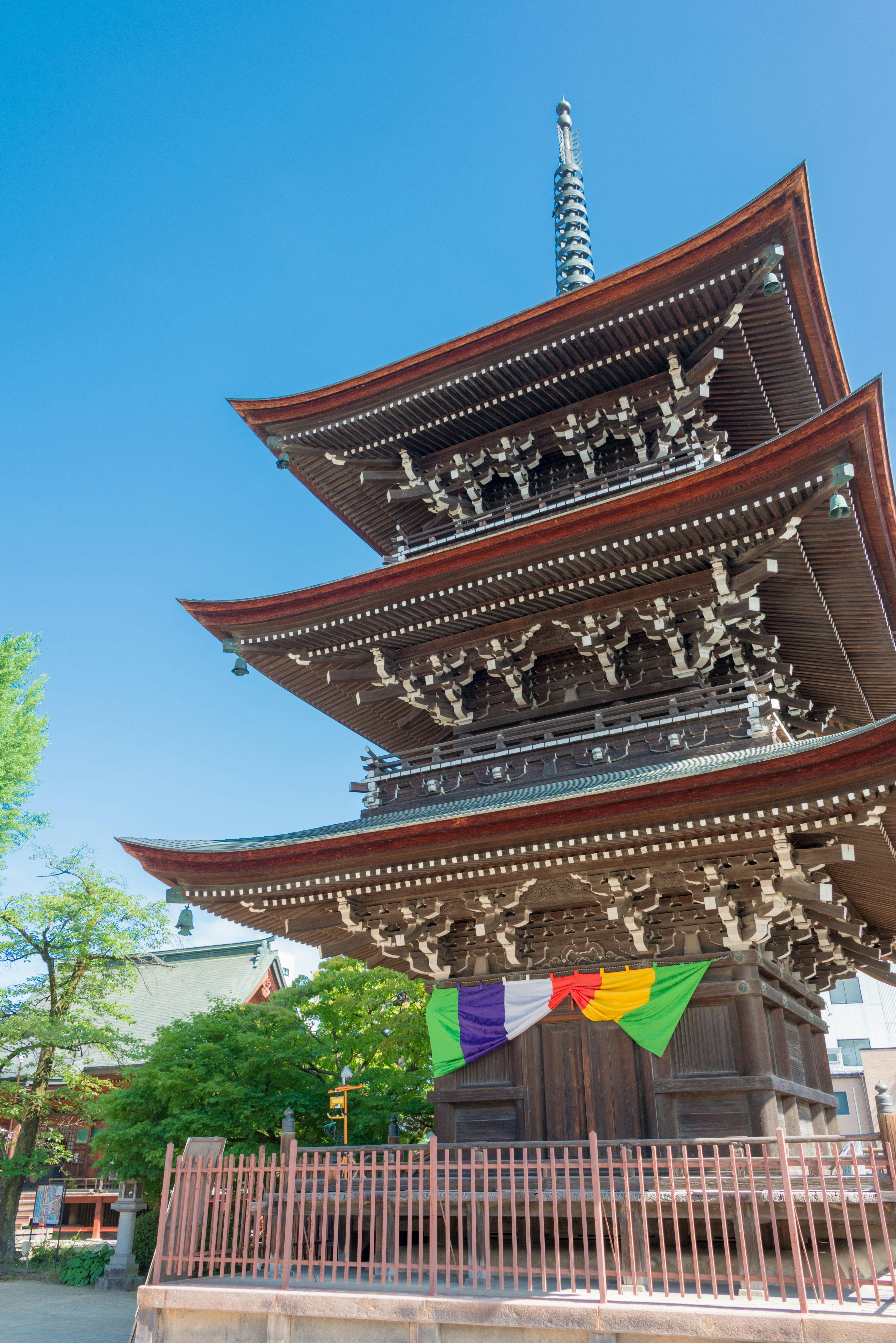 Three-story wooden pagoda with ornate eaves and a tall spire, decorated with colorful hanging banners at the base. The structure is surrounded by a fence, greenery, and set against a clear blue sky.