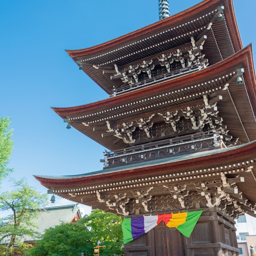 Hida Kokubun-ji Temple Three-story wooden pagoda with ornate eaves and a tall spire, decorated with colorful hanging banners at the base. The structure is surrounded by a fence, greenery, and set against a clear blue sky.