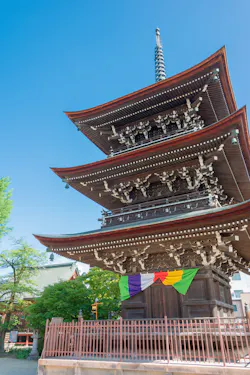 Three-story wooden pagoda with ornate eaves and a tall spire, decorated with colorful hanging banners at the base. The structure is surrounded by a fence, greenery, and set against a clear blue sky.