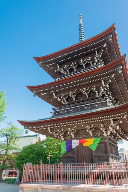 Three-story wooden pagoda with ornate eaves and a tall spire, decorated with colorful hanging banners at the base. The structure is surrounded by a fence, greenery, and set against a clear blue sky.