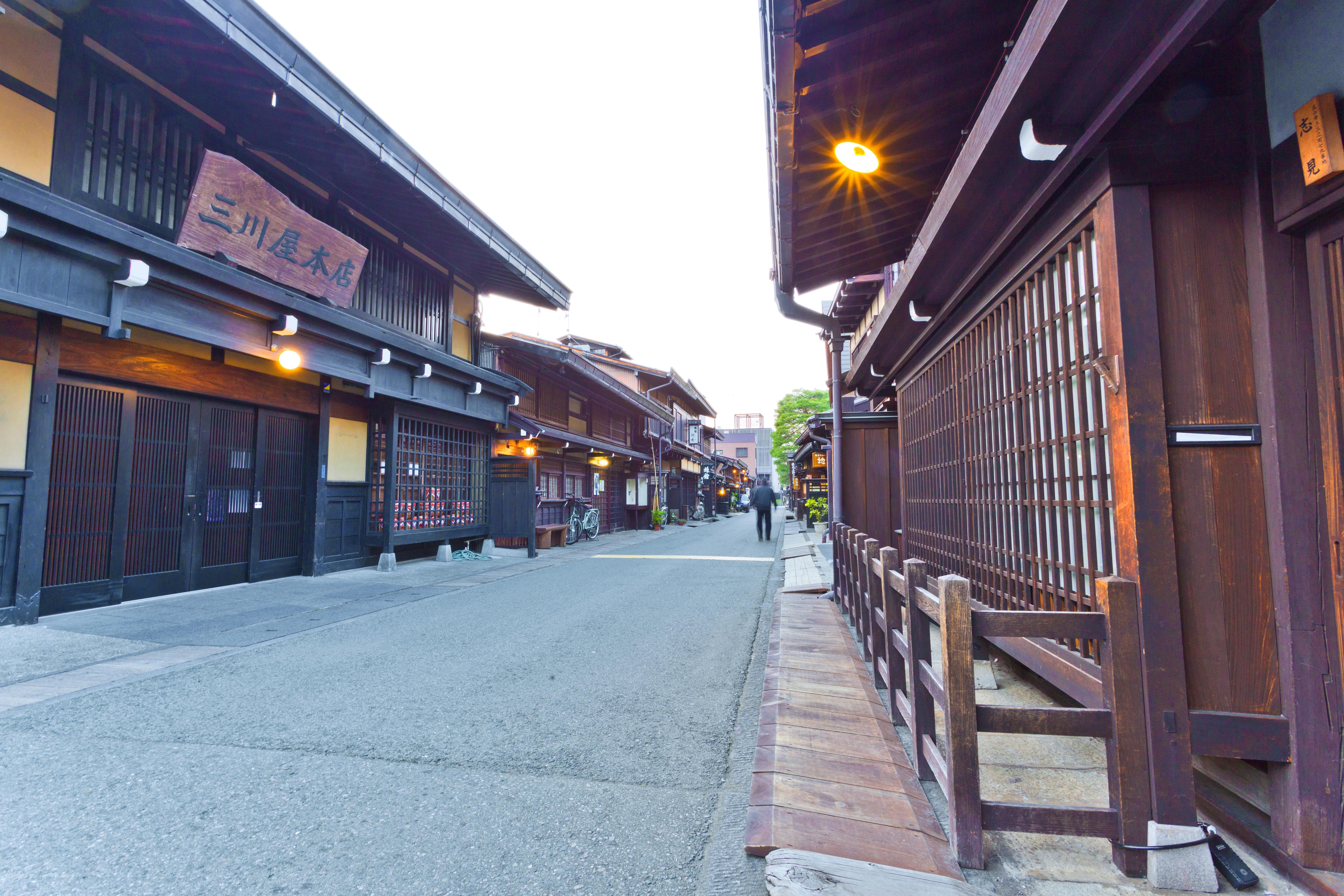 A quiet street lined with traditional wooden buildings in a historic Japanese town, featuring wooden lattice doors and warm lantern lights, with a few people walking in the distance.