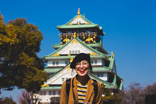 A smiling woman wearing a beret and glasses stands in front of Osaka Castle in Japan, with bright blue sky and trees in the background.