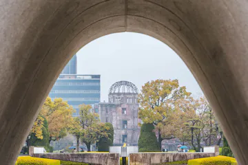 Hiroshima Peace Memorial Park View through the Hiroshima Peace Memorial Park cenotaph arch, with the Atomic Bomb Dome framed in the background, surrounded by trees, reflecting pool, and a stone monument with Japanese inscription in the foreground.