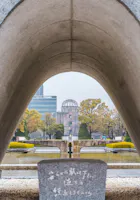 View through the Hiroshima Peace Memorial Park cenotaph arch, with the Atomic Bomb Dome framed in the background, surrounded by trees, reflecting pool, and a stone monument with Japanese inscription in the foreground.