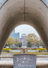 View through the Hiroshima Peace Memorial Park cenotaph arch, with the Atomic Bomb Dome framed in the background, surrounded by trees, reflecting pool, and a stone monument with Japanese inscription in the foreground.