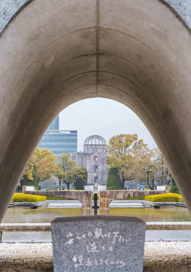 View through the Hiroshima Peace Memorial Park cenotaph arch, with the Atomic Bomb Dome framed in the background, surrounded by trees, reflecting pool, and a stone monument with Japanese inscription in the foreground.