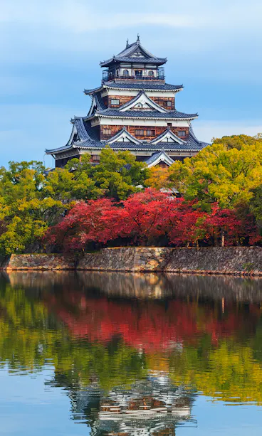 Hiroshima-jo Castle A traditional Japanese castle stands beside a calm moat, surrounded by autumn trees with red, yellow, and green leaves, reflecting on the water under a partly cloudy sky.