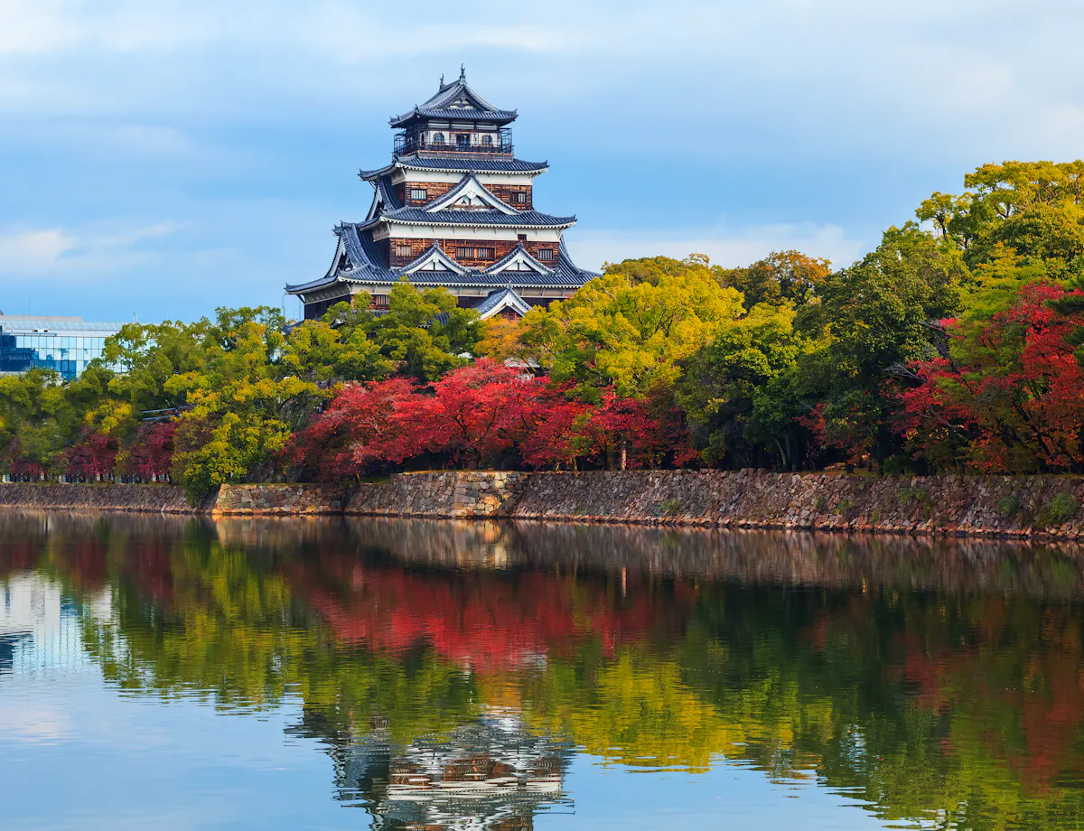 Hiroshima-jo Castle A traditional Japanese castle stands beside a calm moat, surrounded by autumn trees with red, yellow, and green leaves, reflecting on the water under a partly cloudy sky.