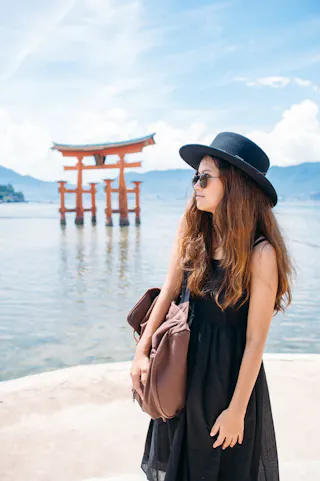 A woman in a black dress and hat stands by the water, holding a bag, with the famous red Torii gate of Itsukushima Shrine in Japan visible in the background under a blue sky.