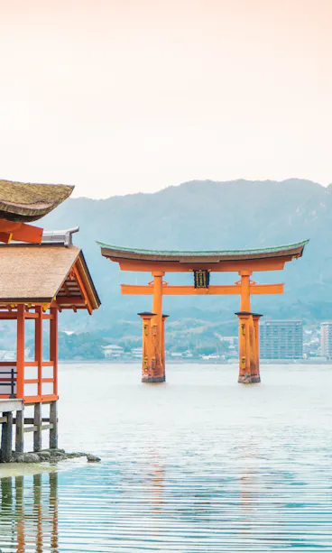 Miyajima A serene view of Itsukushima Shrine in Japan, featuring traditional wooden buildings over water and the iconic large red Torii gate in the sea. Mountains rise in the background under a cloudy sky, while two visitors stand on a deck enjoying the scenery.