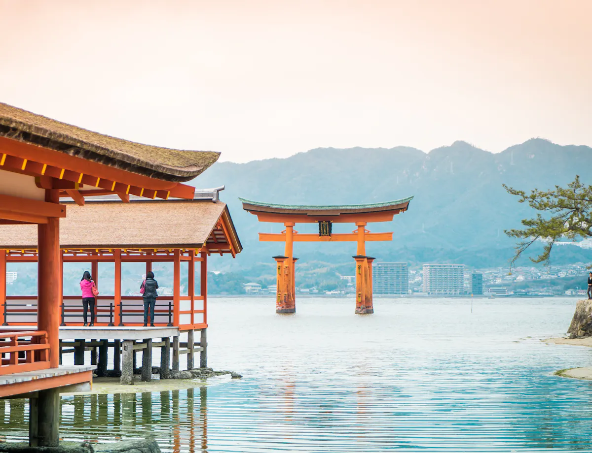 Miyajima A serene view of Itsukushima Shrine in Japan, featuring traditional wooden buildings over water and the iconic large red Torii gate in the sea. Mountains rise in the background under a cloudy sky, while two visitors stand on a deck enjoying the scenery.