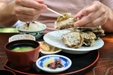 A person uses chopsticks to eat an oyster from a plate of oysters, with bowls of rice, miso soup, pickles, and condiments on a wooden table.