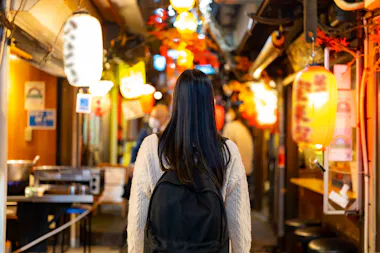 A woman with long dark hair and a black backpack walks through a narrow alley decorated with colorful lanterns and lights, with shops and cafes lining both sides.