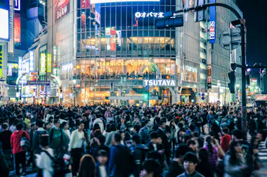 Crowded street scene at night in Shibuya, Tokyo, with people crossing and commercial buildings in the background. Neon signs light up the area, with a Starbucks and Tsutaya prominently visible in the center.