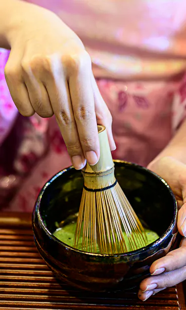 Matcha Making A person wearing a pink kimono whisks matcha green tea in a black bowl using a bamboo whisk, with a small decorated container nearby on a wooden tray.