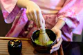 A person wearing a pink kimono whisks matcha green tea in a black bowl using a bamboo whisk, with a small decorated container nearby on a wooden tray.