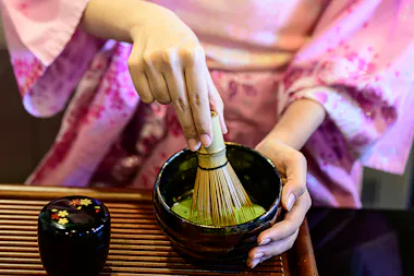 A person wearing a pink kimono whisks matcha green tea in a black bowl using a bamboo whisk, with a small decorated container nearby on a wooden tray.