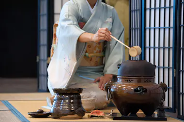 A person in a light blue kimono prepares tea during a traditional Japanese tea ceremony, using a ladle to pour hot water from an iron kettle on a tatami mat floor.