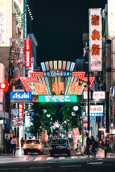 A vibrant street scene in Dotonbori, Osaka at night, with colorful neon signs in Japanese, illuminated shopfronts, cars, bicycles, and people walking along the busy street.
