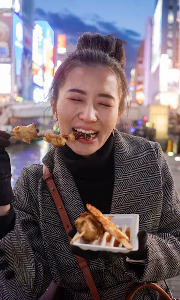 Woman Eating Kushikatsu A woman wearing a coat and gloves smiles with closed eyes while eating street food by a canal at dusk, with colorful city lights and people in the background.