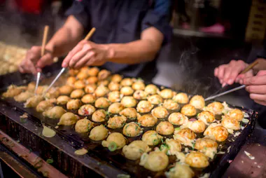 Close-up of hands using chopsticks to cook and turn takoyaki (Japanese octopus balls) on a large griddle, with steam rising and ingredients visible.