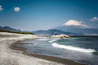 A tranquil beach with dark sand and gentle waves, concrete wave breakers along the shore, and Mount Fuji with a snow-capped peak rising in the distance under a clear blue sky.
