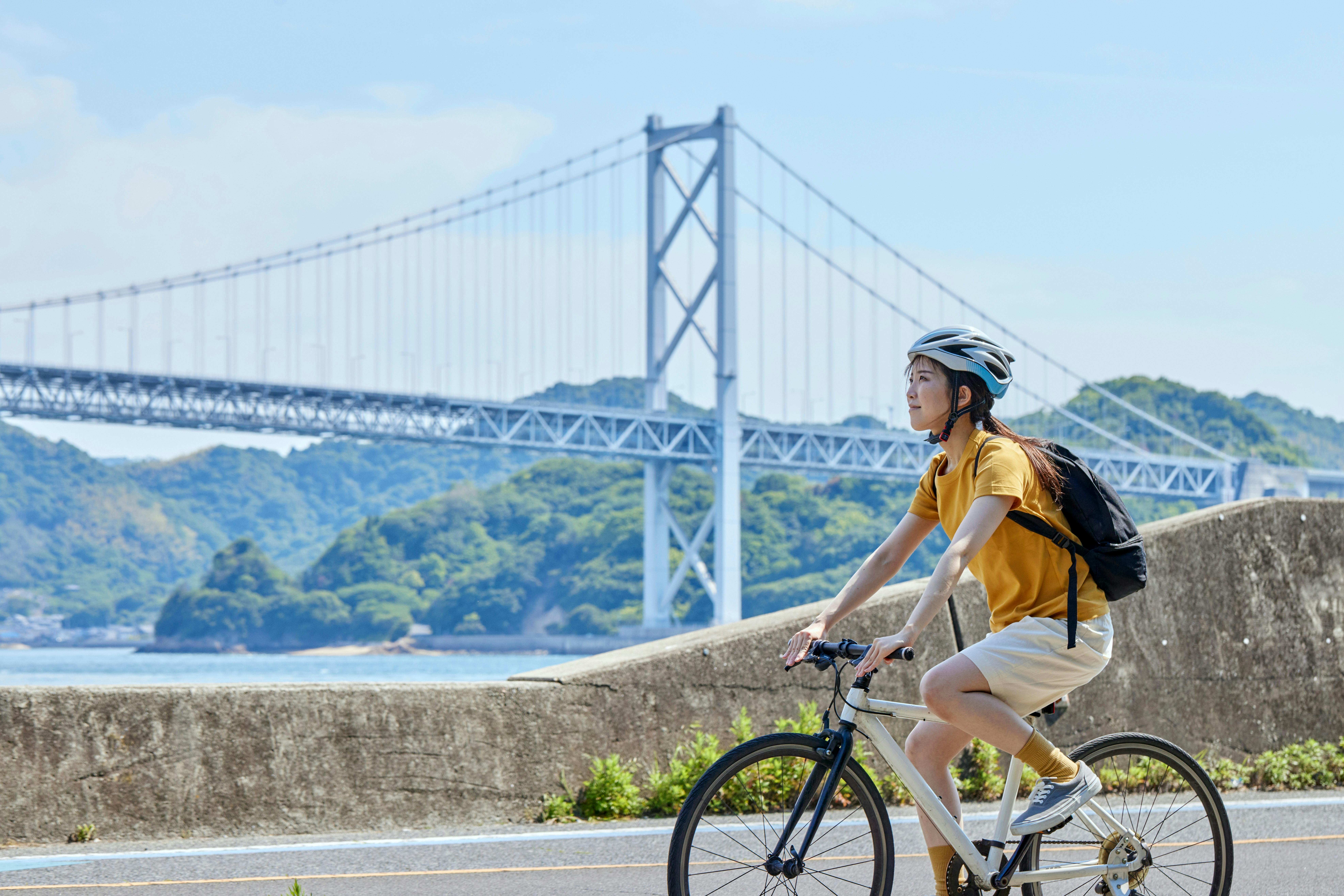 A person wearing a helmet and yellow shirt rides a bicycle along a waterfront path, with a large suspension bridge and green hills in the background under a blue sky.