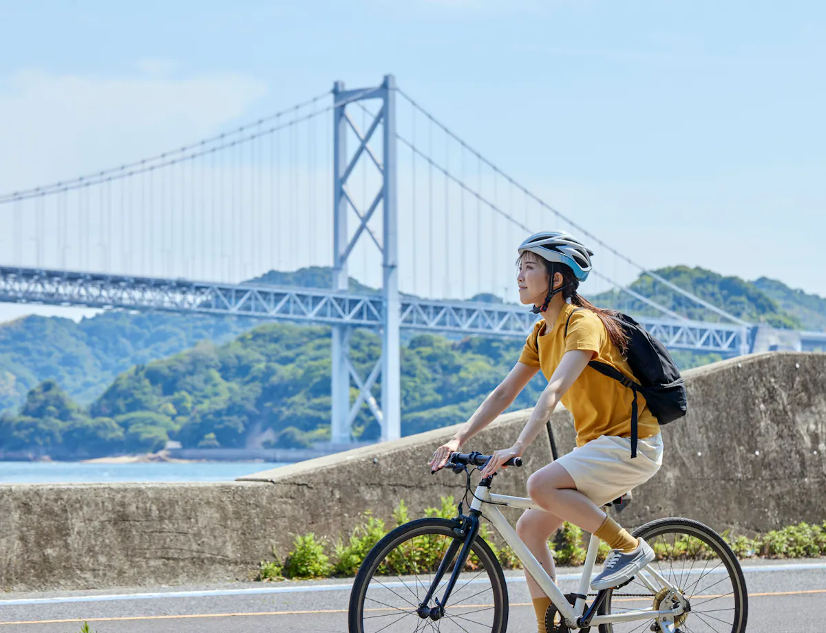 A person wearing a helmet and yellow shirt rides a bicycle along a waterfront path, with a large suspension bridge and green hills in the background under a blue sky.
