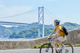 A person wearing a helmet and yellow shirt rides a bicycle along a waterfront path, with a large suspension bridge and green hills in the background under a blue sky.