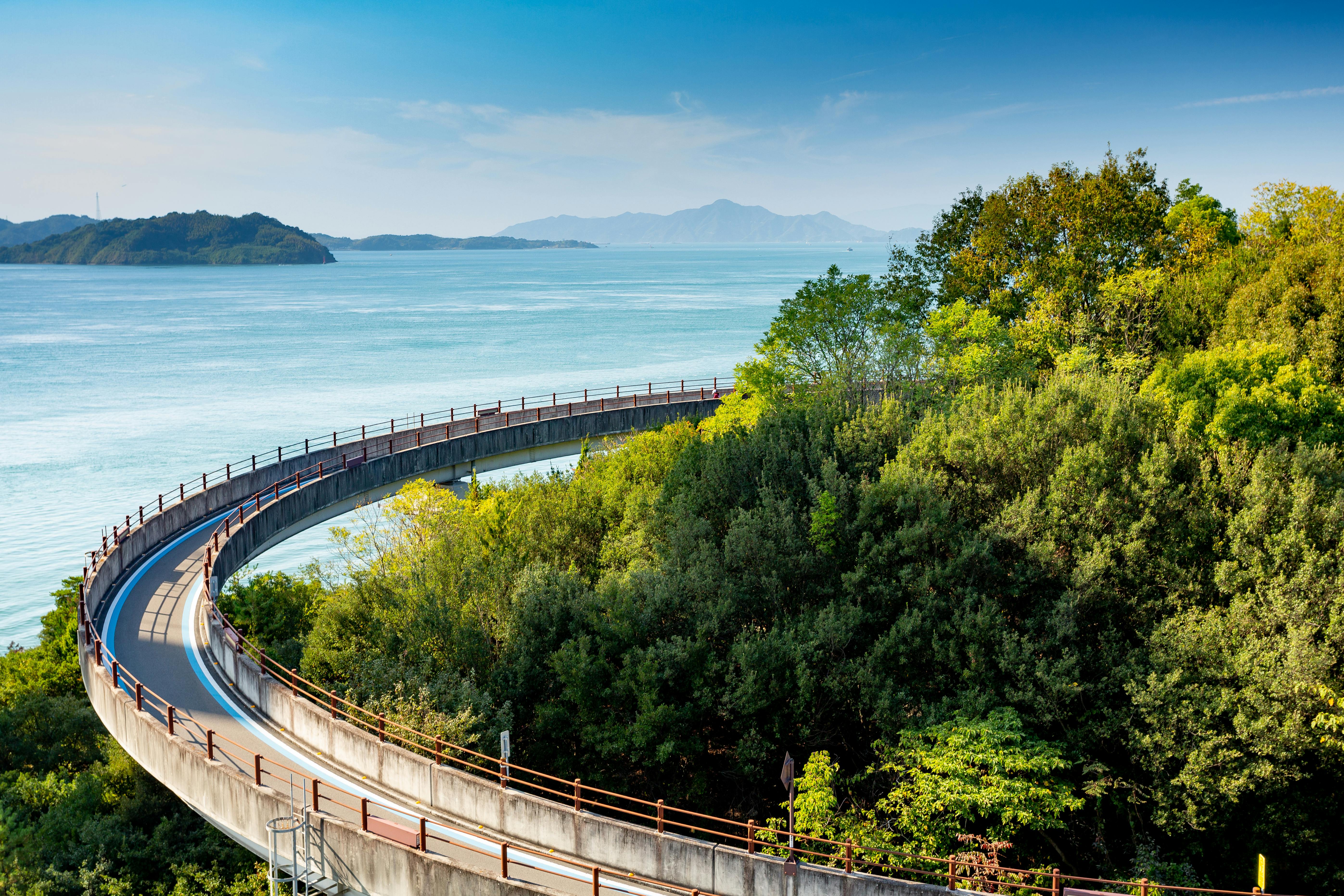 A curved elevated walkway winds through lush green trees beside a calm blue sea, with distant islands and mountains visible under a clear sky.