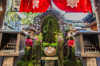A moss-covered Buddhist statue is flanked by two smaller statues wearing red bibs, surrounded by flowers and lanterns, beneath vibrant red banners at a Japanese temple.