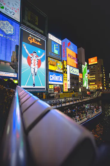 Bright neon billboards, including the famous running Glico Man, light up the Dotonbori district in Osaka, Japan at night. People ride on a boat along the river below the illuminated signs.