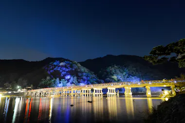 A long, illuminated wooden bridge stretches across a calm river at night, with tree-covered mountains in the background and colorful lights reflecting on the water.