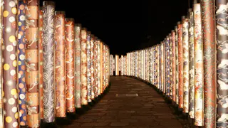 A stone pathway is lined on both sides by tall, illuminated cylindrical pillars decorated with colorful, intricate patterns, creating a glowing tunnel effect at night.