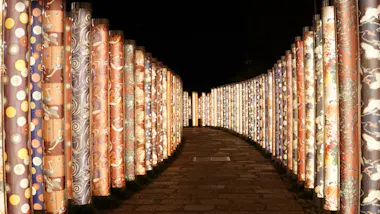 A stone pathway is lined on both sides by tall, illuminated cylindrical pillars decorated with colorful, intricate patterns, creating a glowing tunnel effect at night.