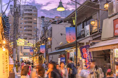 A bustling street market in Japan at dusk, with blurred crowds walking among old-style shops and colorful signs. Warm lights illuminate the scene, and taller modern buildings rise in the background under a purple sky.