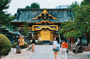 Several people walk along a stone path toward an ornate, gold-trimmed Japanese temple surrounded by greenery and traditional statues on a bright day.