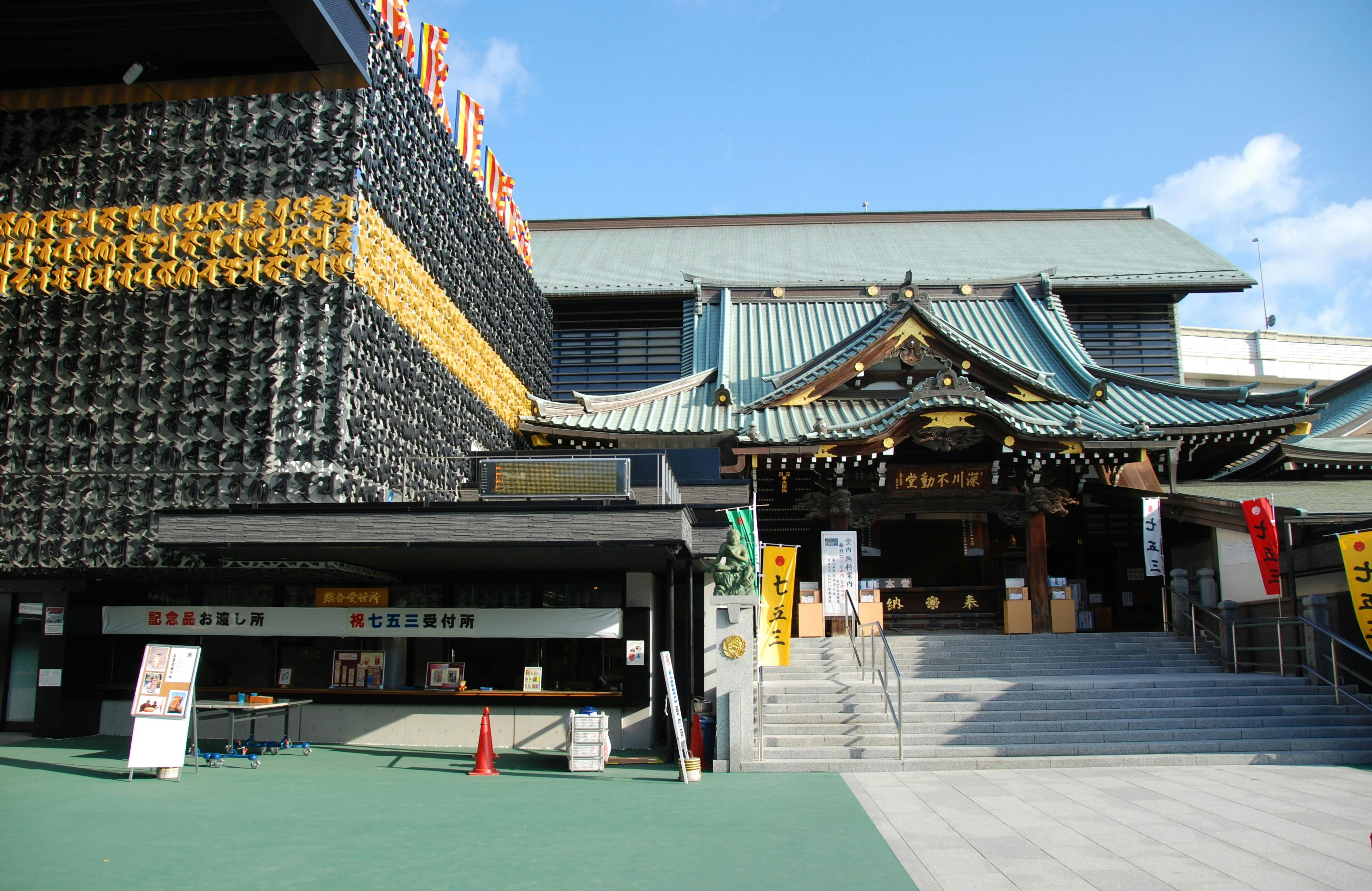 The image shows the exterior of a traditional Japanese shrine with a modern black building adjacent to it. The shrine has a green, ornate roof and wooden details, while the contemporary building features a geometric facade covered in patterns. A set of stairs leads to the shrine.