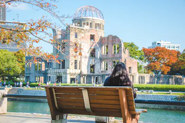 A woman with long dark hair sits on a wooden bench facing the Hiroshima Peace Memorial, also known as the Atomic Bomb Dome, on a sunny day with autumn foliage and a clear blue sky.