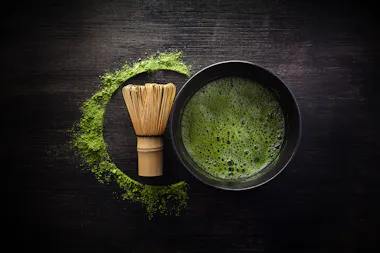 A bamboo matcha whisk and a black bowl of frothy matcha tea sit on a dark wooden surface, with green matcha powder artfully arranged in a semi-circle around the whisk and bowl.