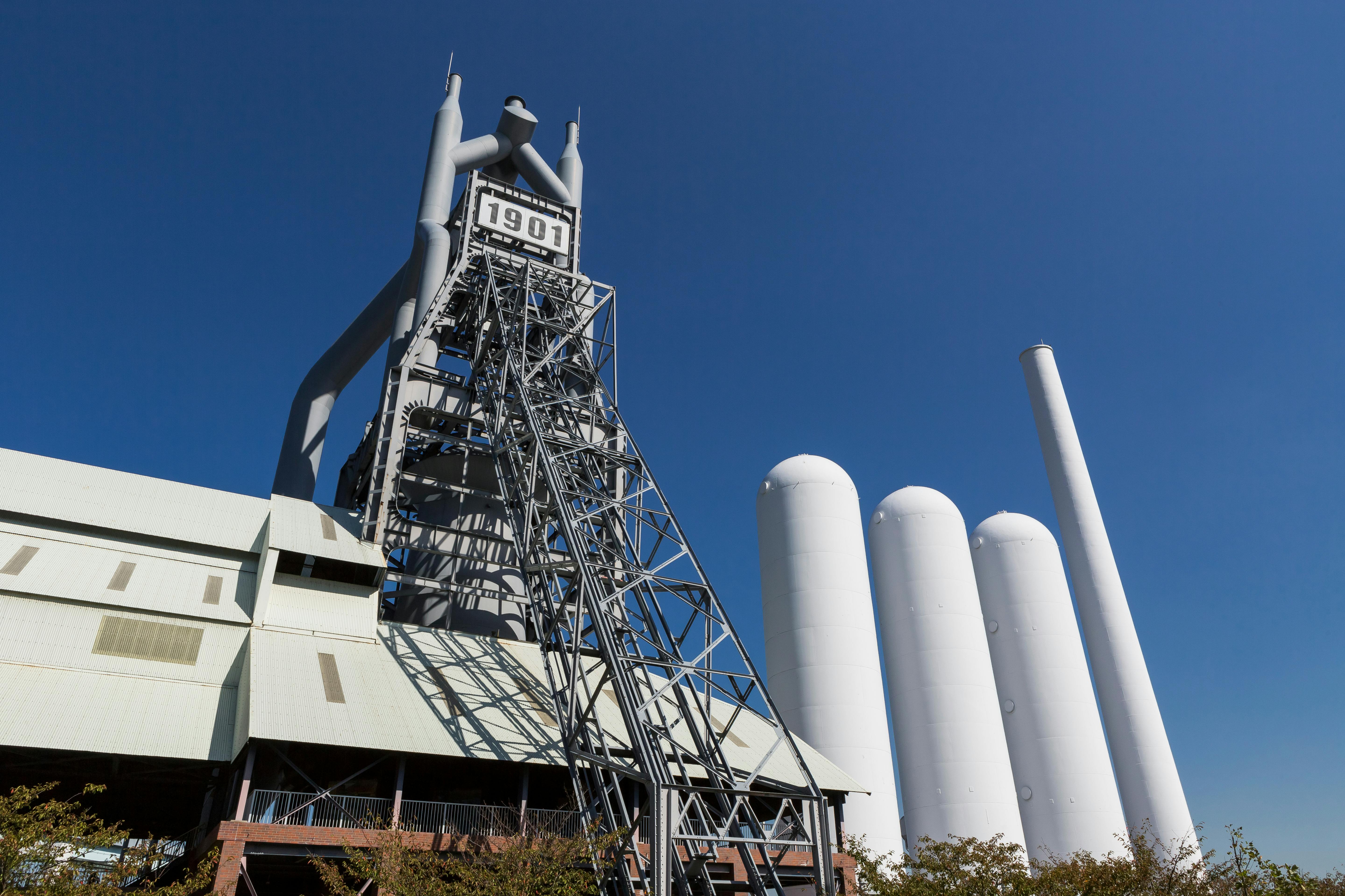 A large industrial structure with "1901" displayed at the top, tall white cylindrical tanks, and a slanted metal framework, set against a clear blue sky.
