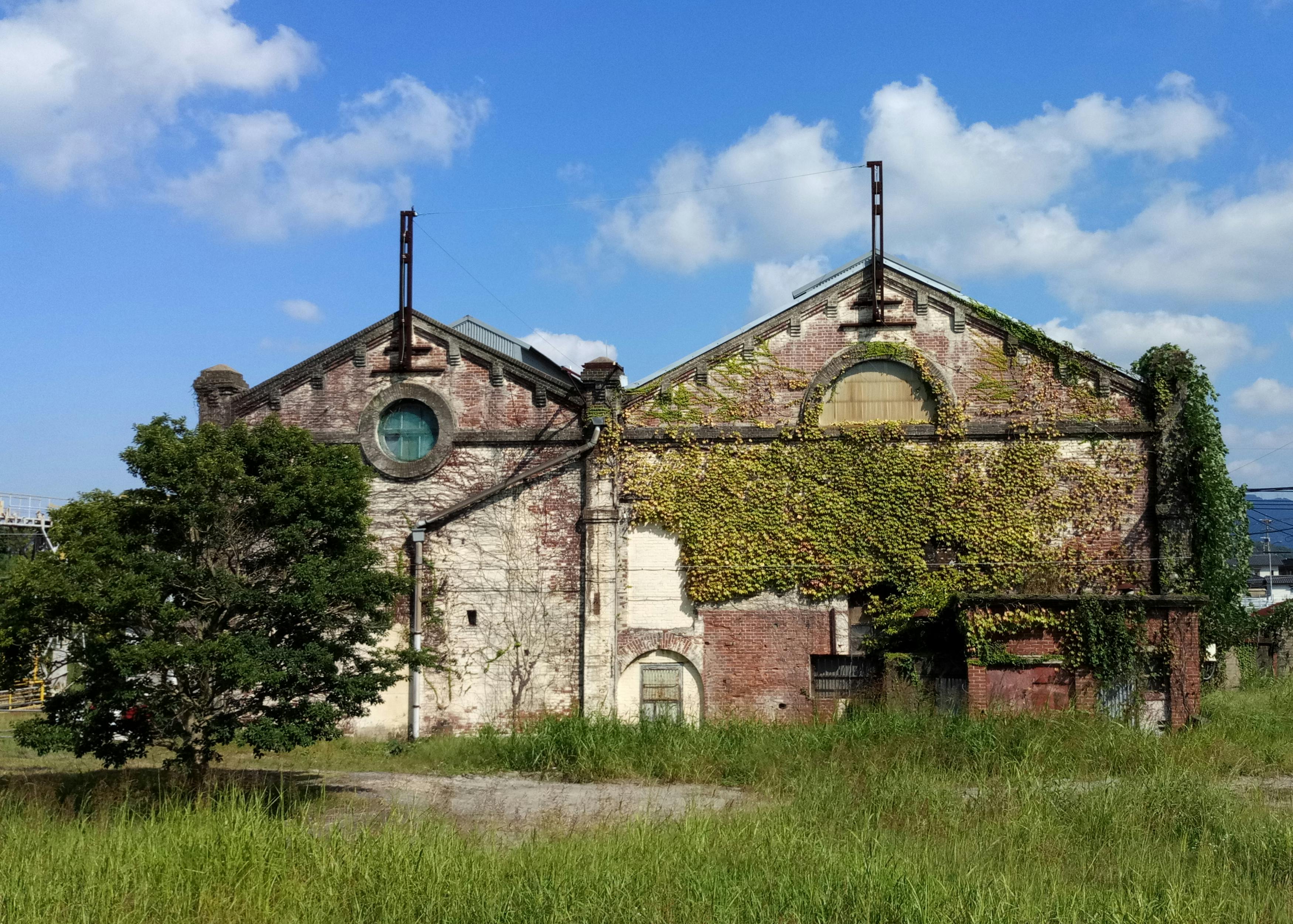 An old, brick industrial building with ivy covering part of its facade, circular and arched windows, and surrounded by overgrown grass and a tree, under a blue sky with scattered clouds.