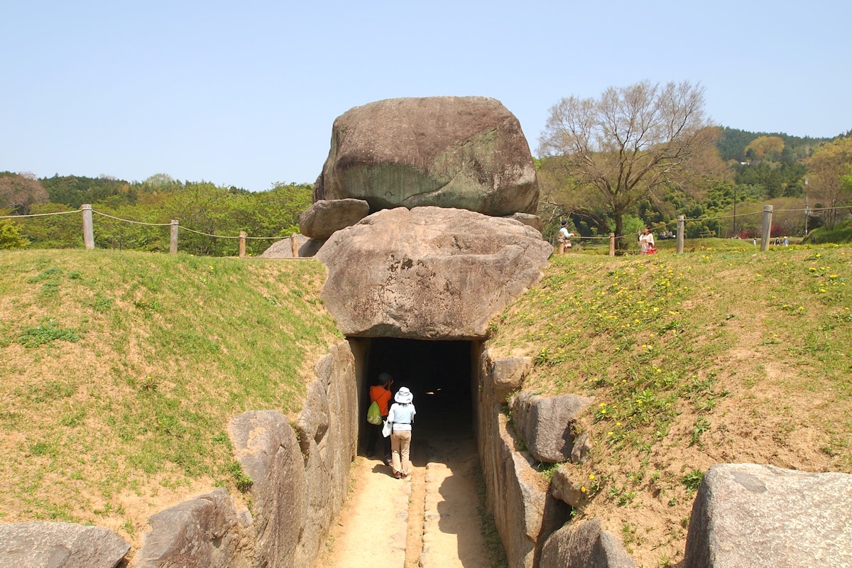 Ishibutai Tomb A stone-lined pathway leads to the entrance of an ancient rock tomb, with large boulders forming its structure. Several visitors are entering the tomb. Green grass and a few leafless trees surround the site under a clear blue sky.