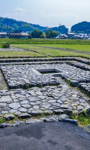 Itabuki Imperial Palace Ruins Ancient stone foundations in a square pattern on grassy land, with houses and trees in the background under a partly cloudy sky.