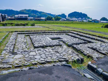 Ancient stone foundations in a square pattern on grassy land, with houses and trees in the background under a partly cloudy sky.