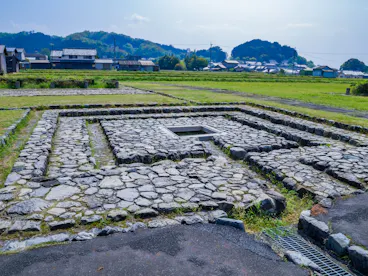 Ancient stone foundations in a square pattern on grassy land, with houses and trees in the background under a partly cloudy sky.
