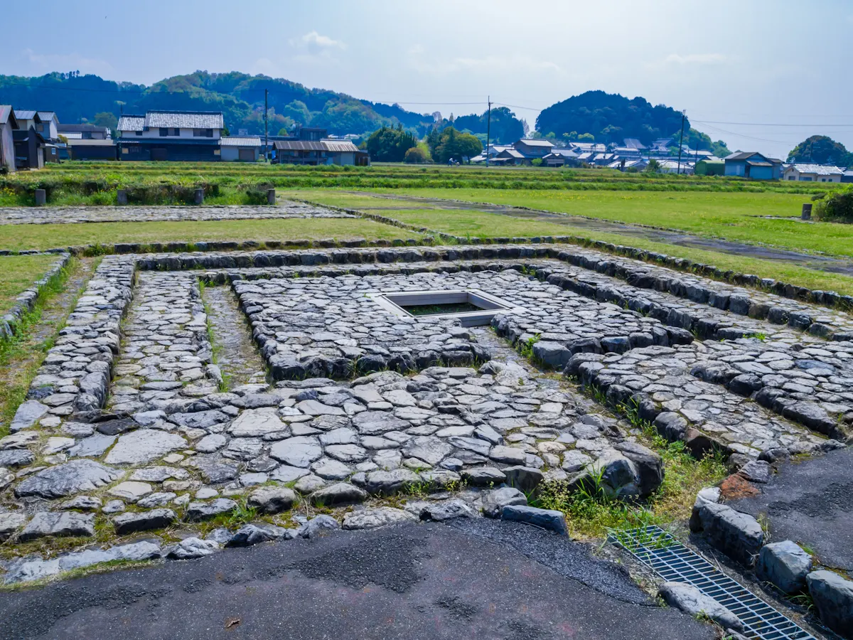 Ancient stone foundations in a square pattern on grassy land, with houses and trees in the background under a partly cloudy sky.
