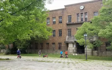 Four people with bicycles stand in front of an old, brown brick building with a clock above the entrance, surrounded by tall green trees and a cobblestone courtyard.