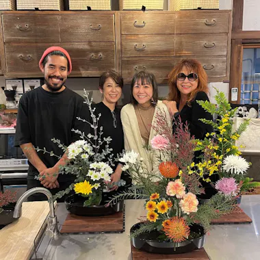 Kado Four people stand smiling behind a table with colorful floral arrangements in a cozy room with wooden cabinets and kitchen appliances in the background.